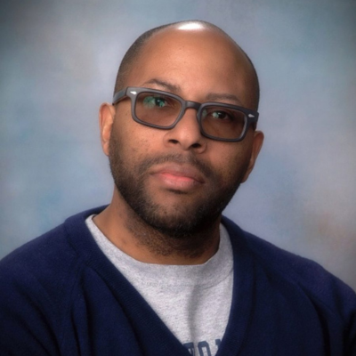 A studio portrait of a man with a shaved head and short facial hair, wearing rectangular glasses with dark frames. He is dressed in a navy blue sweater over a gray shirt. The background is softly blurred in neutral tones, and he faces the camera with a calm, neutral expression.